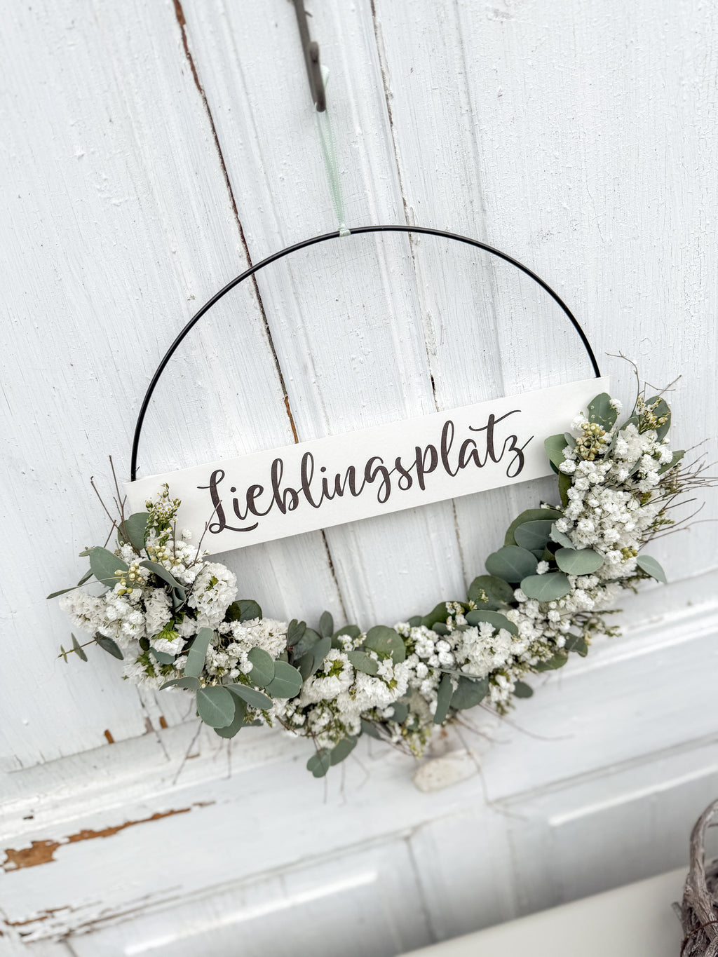 Halb gebundener Metallring mit weißen Trockenblumen und Eukalyptus, dekoriert vor rustikaler Holztüre mit Banner „Lieblingsplatz“.