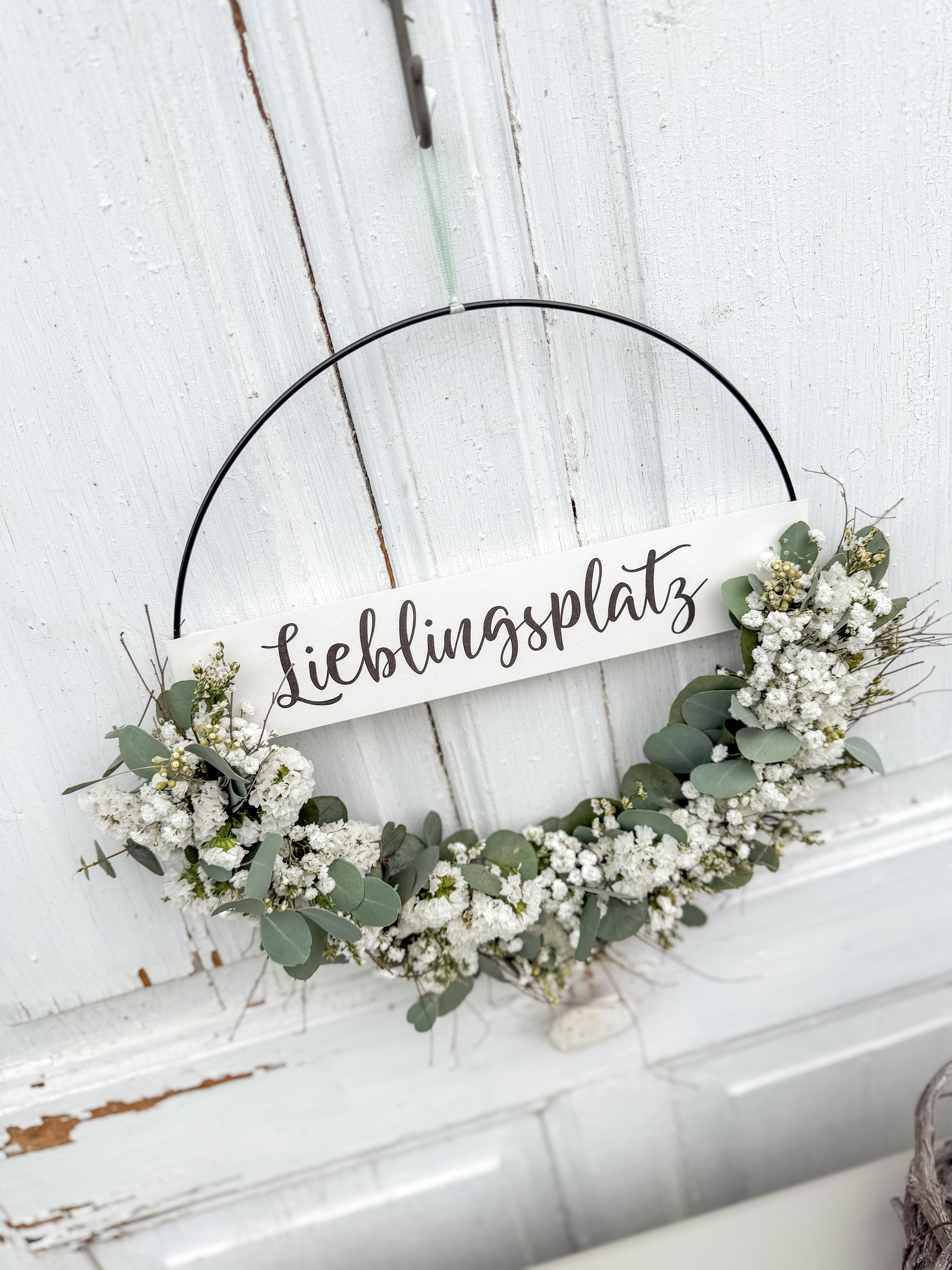 Halb gebundener Metallring mit weißen Trockenblumen und Eukalyptus, dekoriert vor rustikaler Holztüre mit Banner „Lieblingsplatz“.