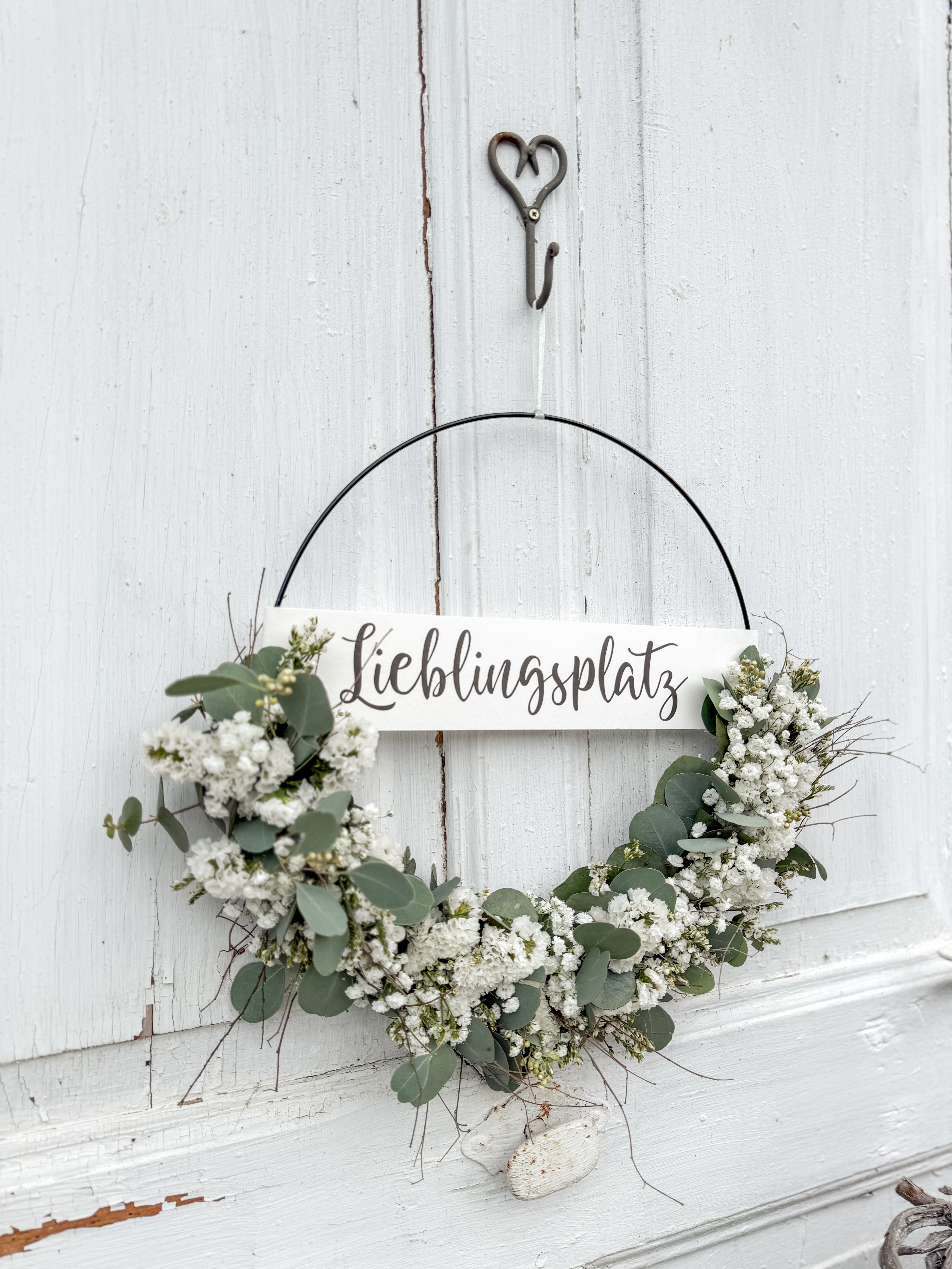 Handgefertigter FlowerLoop „Lieblingsplatz“ mit Eukalyptus und weißen Blüten an einer natürlichen Holztüre dekoriert.