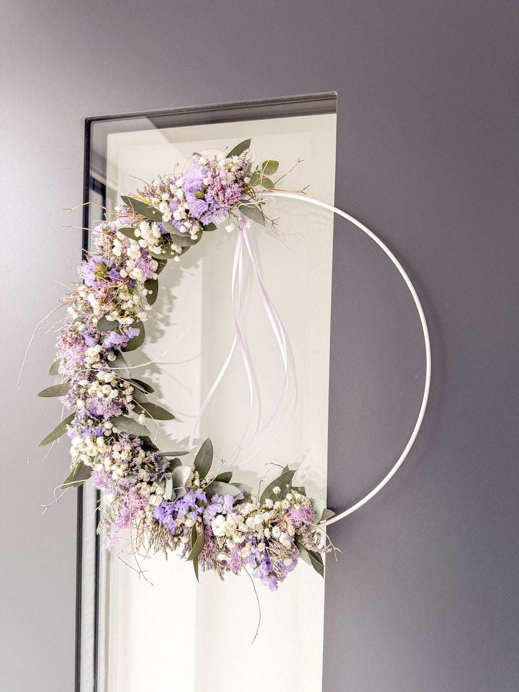 1/2 Loop »Vertical Lila Spring« – Metallring vertikal links bestückt mit Eukalyptus, lila Strandflieder, Limonium, Schleierkraut, Islandmoos in Fliederlila, abgerundet durch Satinband - Bild zeigt Werkstück an dunkler Haustüre