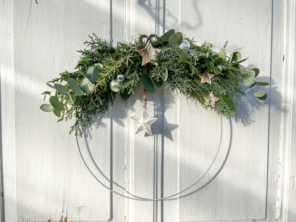 1/2 Winter-Loop ‚Evergreen Canopy‘ Querformat, Metallring halbseitig dekoriert mit Eukalyptus- und Koniferengrün, Zweige überragen oberen Rand, Lerchenzapfen, Birkensterne, Dekokugeln