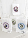 Mehrere Geschenktüten »Mini Flor« nebeneinander, jede dekoriert mit konserviertem Mooskranz und Mini-Bouquet, in verschiedenen Farbvarianten wie Salbei, Bleu und Flieder, handgefertigte Geschenkideen
