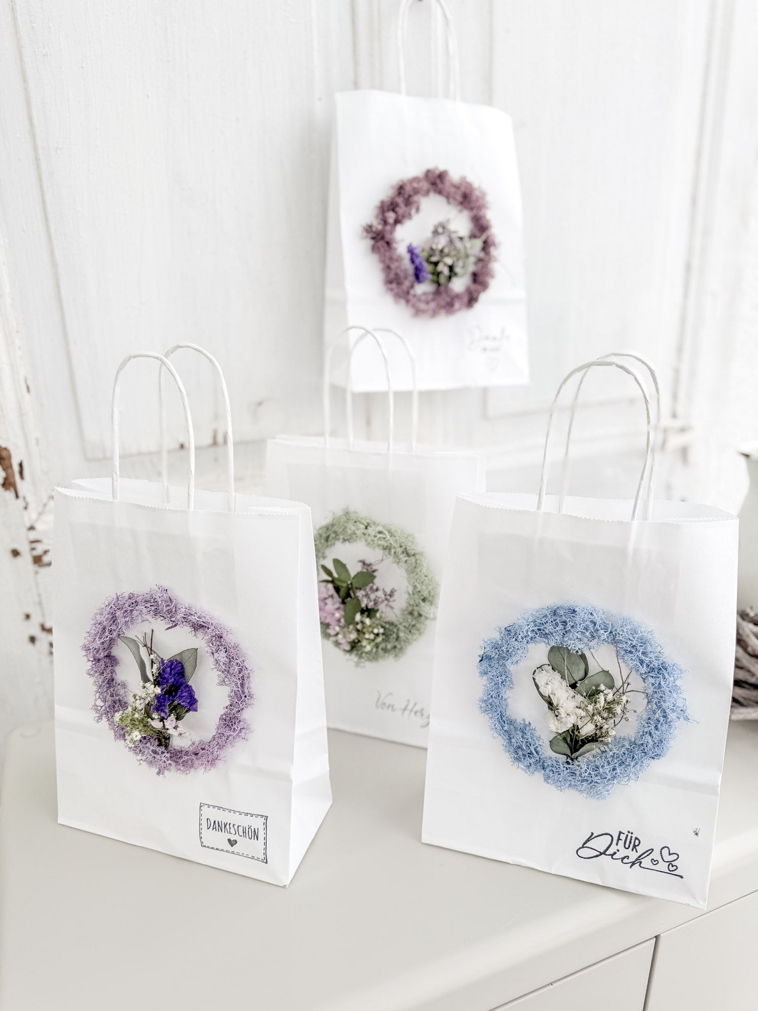 Mehrere Geschenktüten »Mini Flor« nebeneinander, jede dekoriert mit konserviertem Mooskranz und Mini-Bouquet, in verschiedenen Farbvarianten wie Salbei, Bleu und Flieder, handgefertigte Geschenkideen