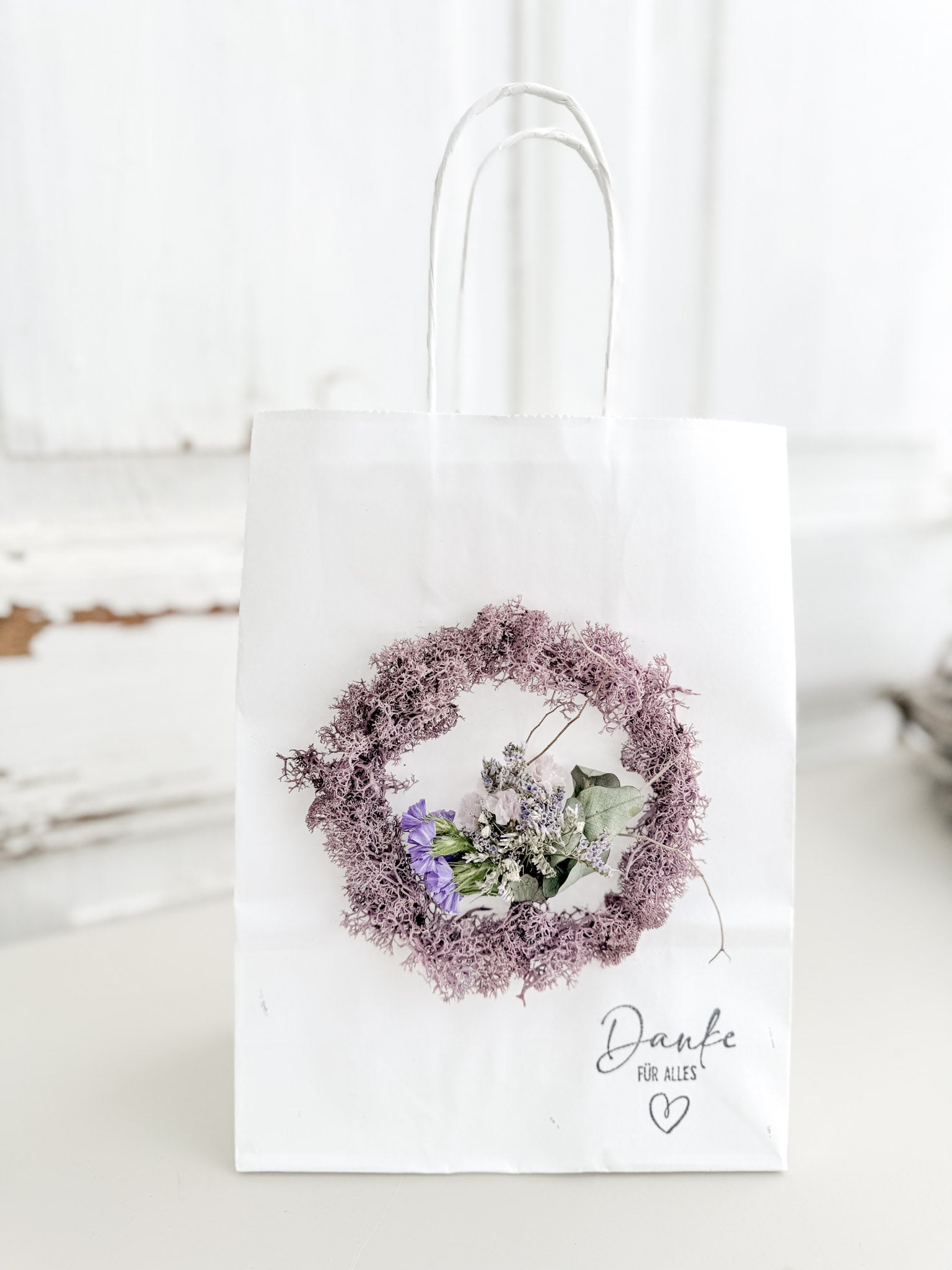 Weiße Geschenktüte »Mini Flor« mit aufgeklebtem Mini-Kranz aus konserviertem Islandmoos in hellem Lila und mittigem Mini-Bouquet aus farblich passenden Blüten