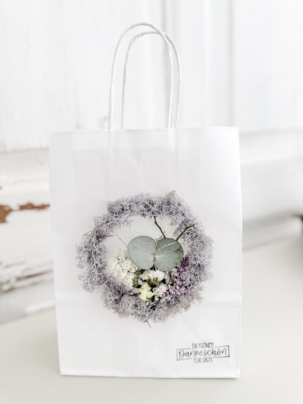 Weiße Geschenktüte »Mini Flor« mit aufgeklebtem Mini-Kranz aus konserviertem Islandmoos in hellem Grau und mittigem Mini-Bouquet aus farblich passenden Blüten