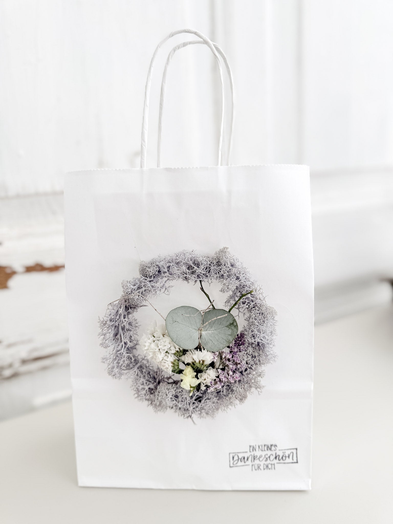 Weiße Geschenktüte »Mini Flor« mit aufgeklebtem Mini-Kranz aus konserviertem Islandmoos in hellem Grau und mittigem Mini-Bouquet aus farblich passenden Blüten