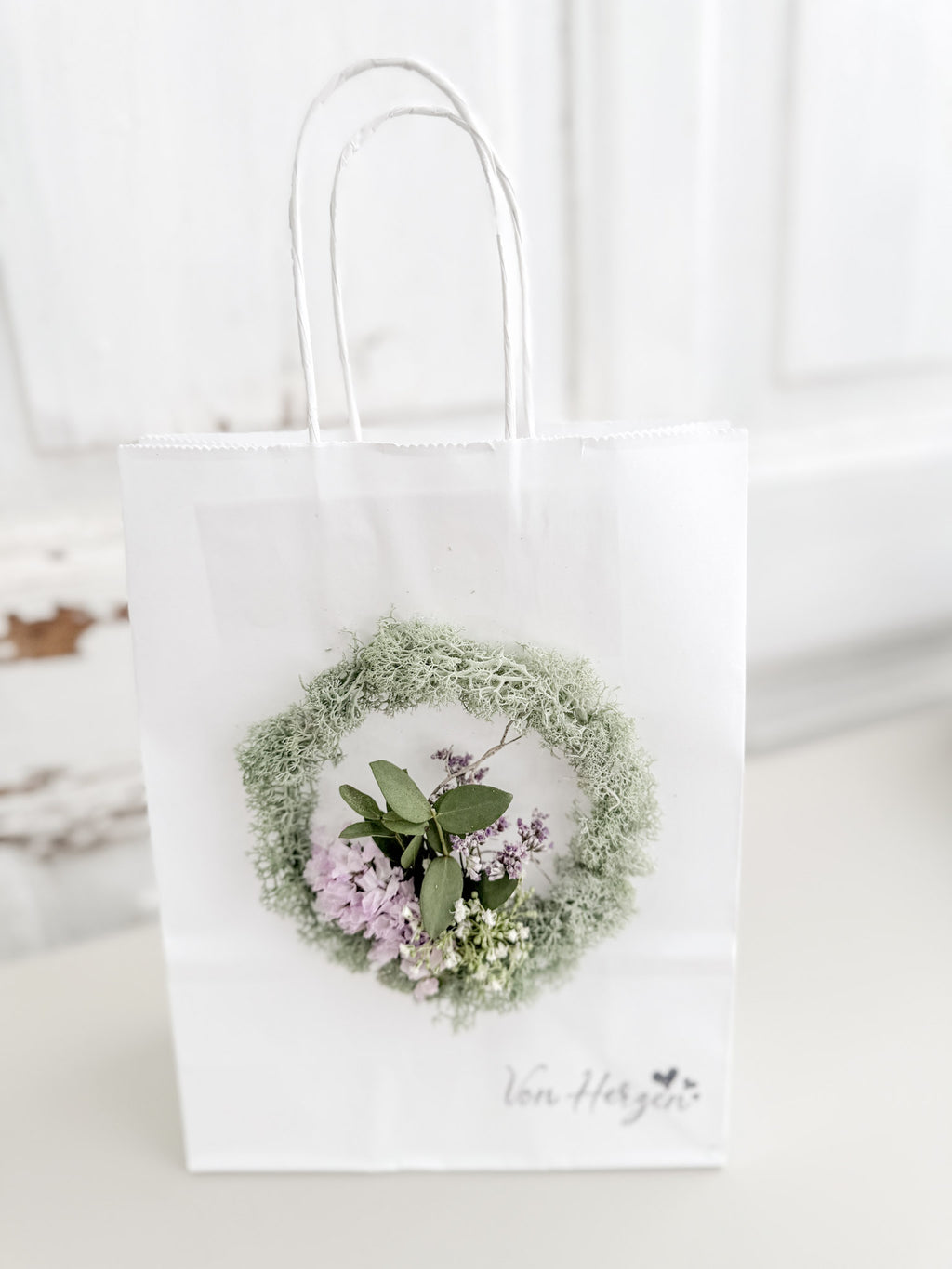 Weiße Geschenktüte »Mini Flor« mit aufgeklebtem Mini-Kranz aus konserviertem Salbeimoos und mittigem Mini-Bouquet aus farblich passenden Blüten