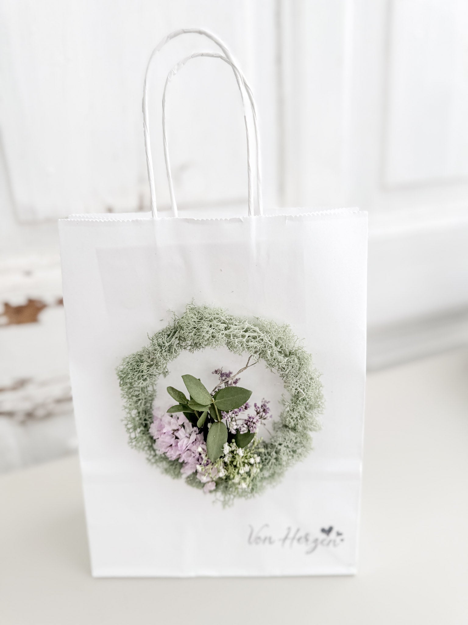 Weiße Geschenktüte »Mini Flor« mit aufgeklebtem Mini-Kranz aus konserviertem Salbeimoos und mittigem Mini-Bouquet aus farblich passenden Blüten