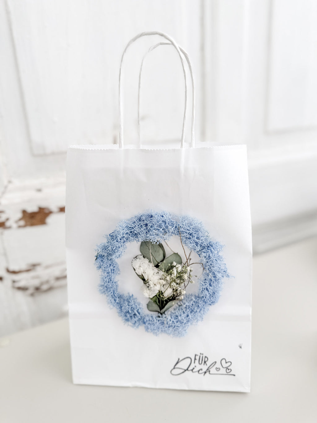 Weiße Geschenktüte »Mini Flor« mit aufgeklebtem Mini-Kranz aus konserviertem Islandmoos in hellem Bleu und mittigem Mini-Bouquet aus farblich passenden Blüten
