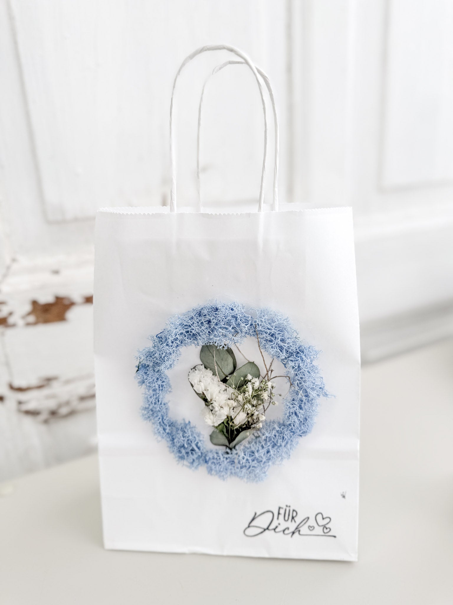Weiße Geschenktüte »Mini Flor« mit aufgeklebtem Mini-Kranz aus konserviertem Islandmoos in hellem Bleu und mittigem Mini-Bouquet aus farblich passenden Blüten