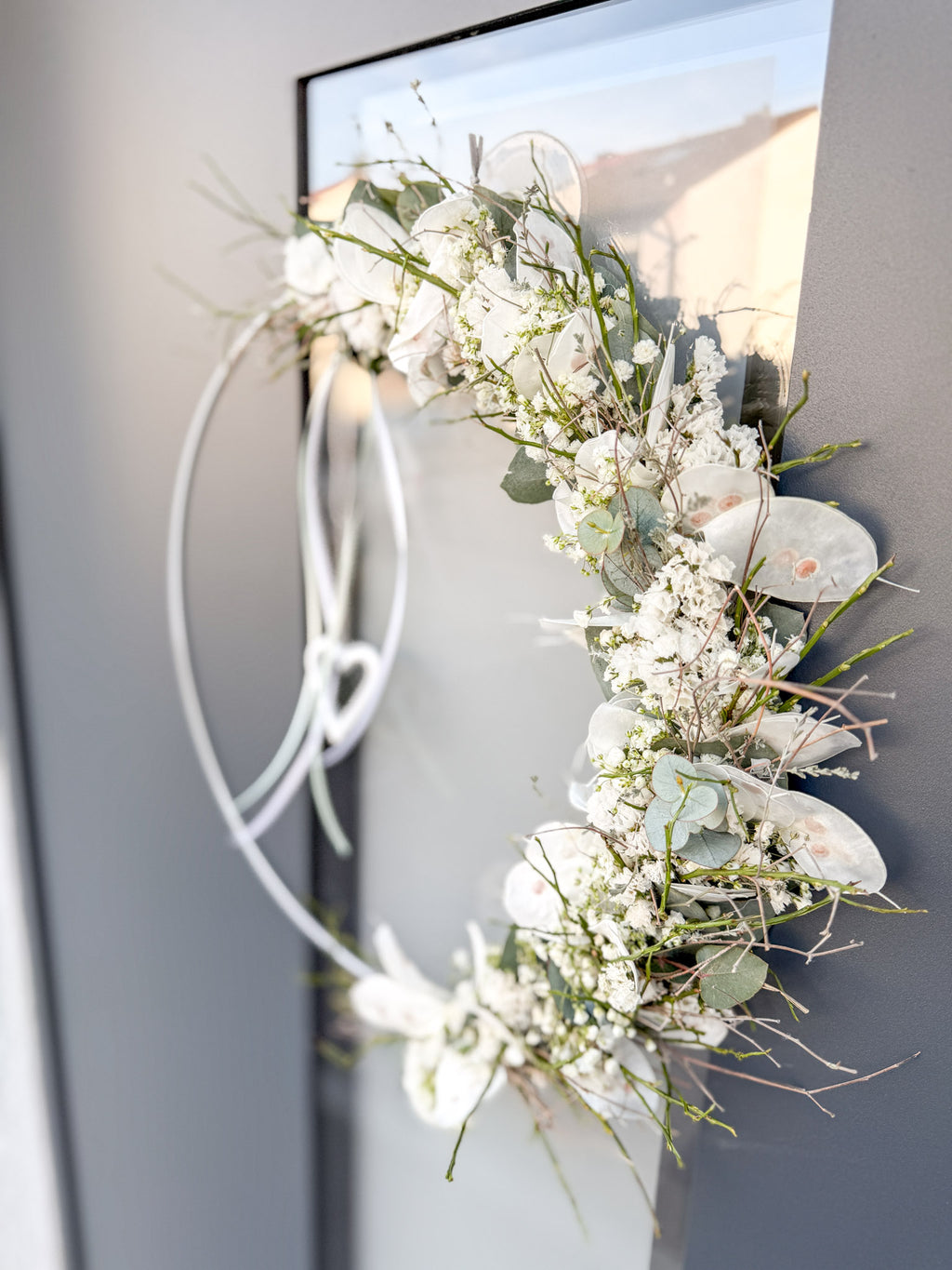 1/2 Loop »Vertical Winterwhite«, vertikal gebundener Metallring mit Eukalyptus, Heidelbeergrün, Schleierkraut und weißen Winterblüten, Satinbänder und hängendes Metallherz - hängend an dunkler Haustüre