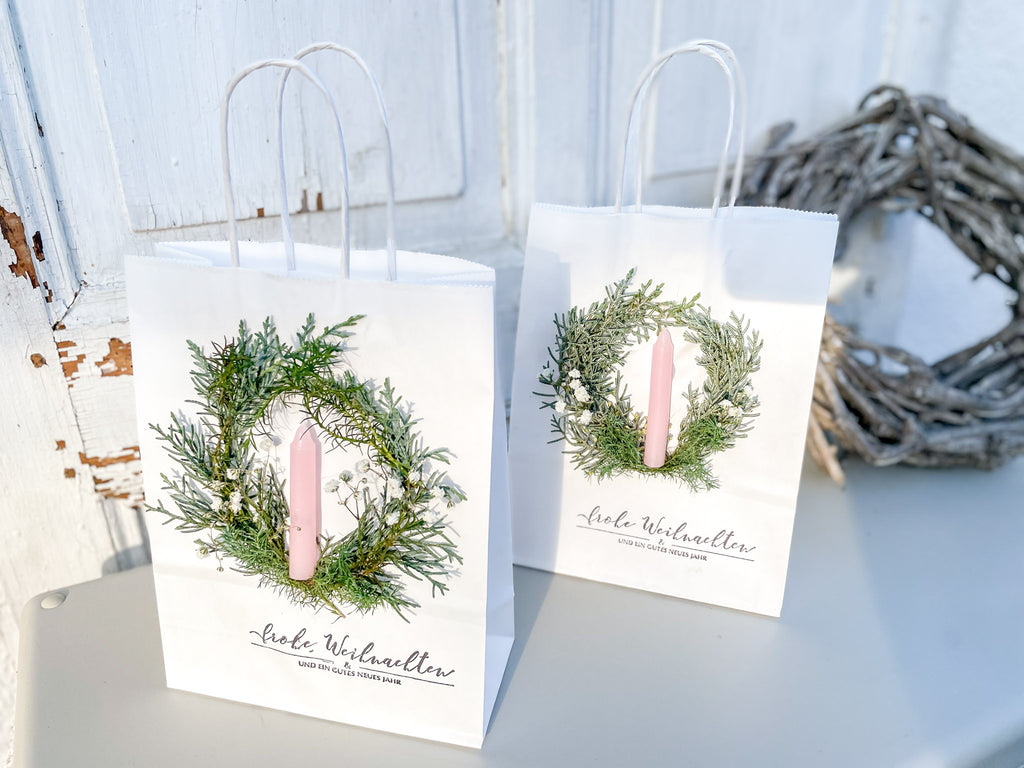 Bild im Querformat, weiße Geschenktüte mit winterlichem Mini-Kranz aus Koniferengrün und Schleierkraut, hellrosa Stabkerze und Stempeldruck „Frohe Weihnachten & ein gutes neues Jahr“, handdekoriert