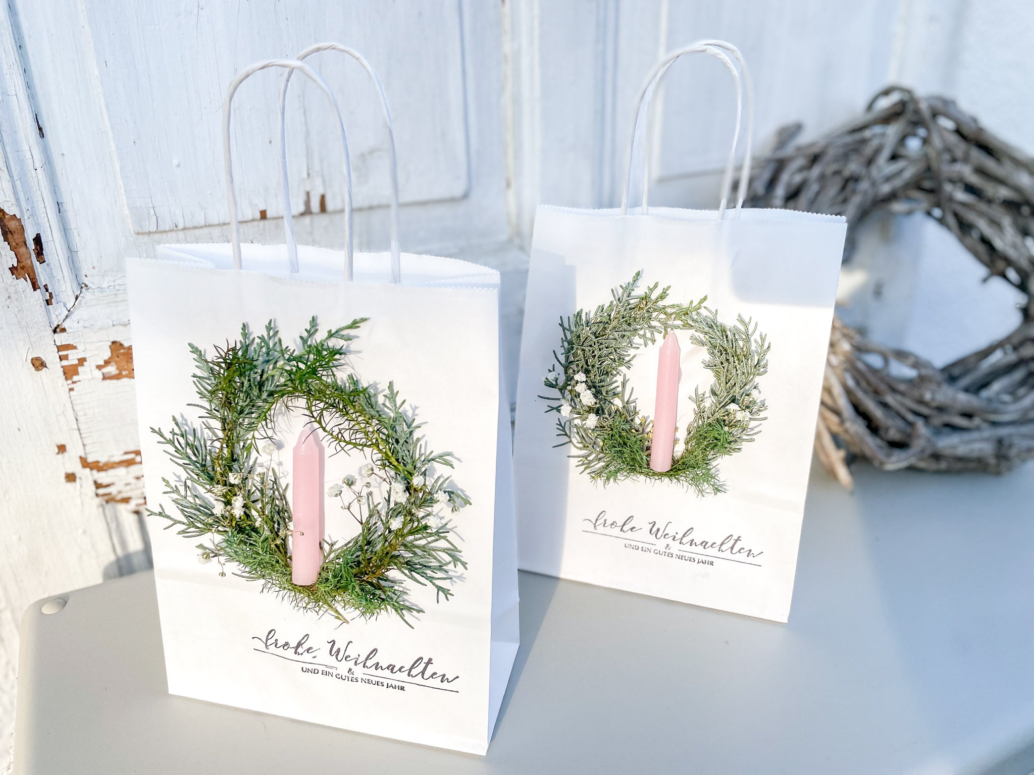 Bild im Querformat, weiße Geschenktüte mit winterlichem Mini-Kranz aus Koniferengrün und Schleierkraut, hellrosa Stabkerze und Stempeldruck „Frohe Weihnachten & ein gutes neues Jahr“, handdekoriert