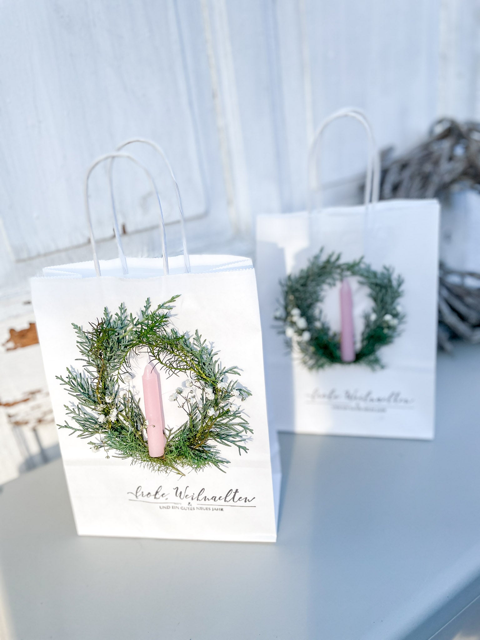 Weiße Geschenktüten mit winterlichem Mini-Kranz aus Koniferengrün und weißem Schleierkraut, hellrosa Stabkerze und Stempeldruck „Frohe Weihnachten & ein gutes neues Jahr“, handdekoriert