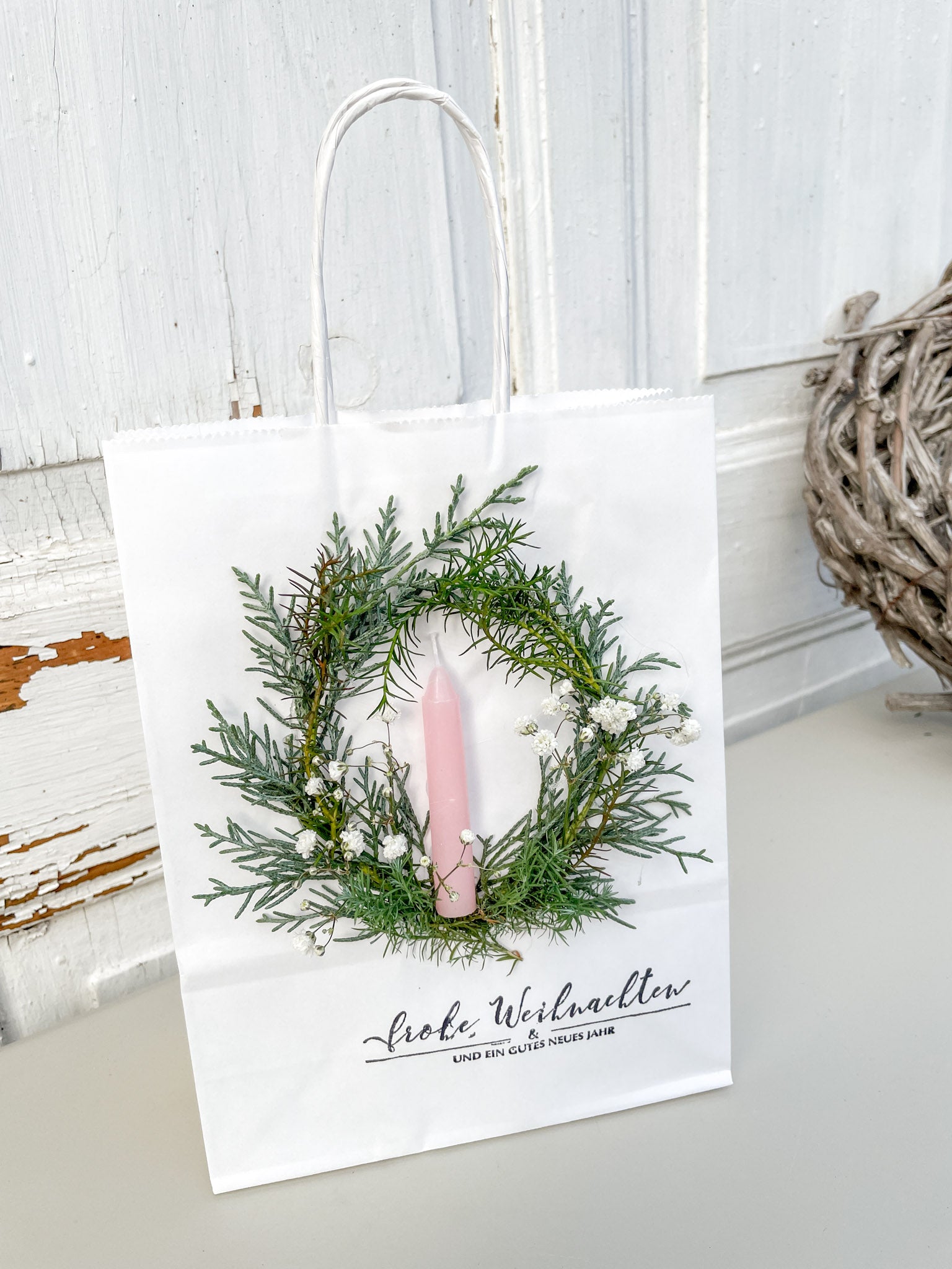 Winterlicher Mini-Kranz aus Koniferengrün und weißem Schleierkraut mit hellrosa Stabkerze auf weißer Geschenktüte, weihnachtliche Geschenkidee