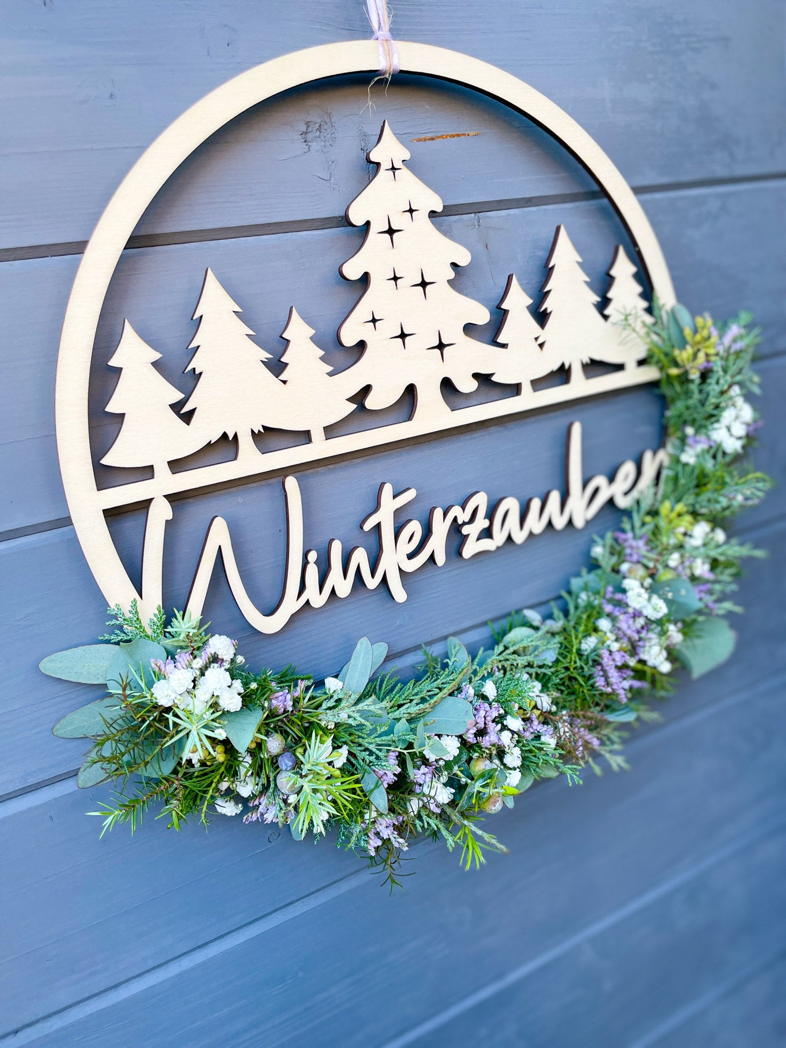 Holzloop Winterzauber mit Schriftzug, bestückt mit Koniferengrün, Eukalyptus und Strandflieder Limonium in Lila, winterliche Dekoration, Ø 25–30 cm, vor grauer Wand