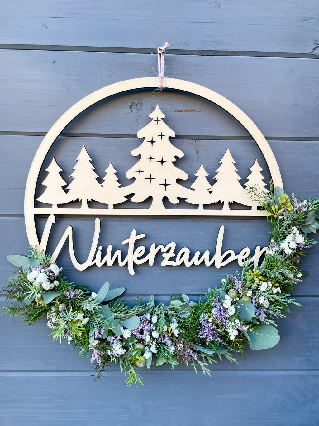 Holzloop Winterzauber mit Schriftzug, bestückt mit Koniferengrün, Eukalyptus und Strandflieder Limonium in Lila, winterliche Dekoration, Ø 25–30 cm, vor grauer Wand