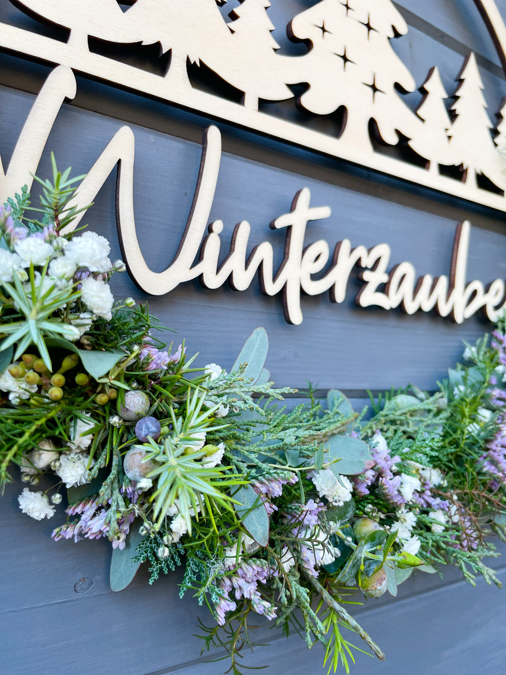 Nahaufnahme Holzloop Winterzauber, Holzschriftzug, Koniferengrün, Eukalyptus, Strandflieder Limonium in Lila, winterliche Dekoration, vor grauer Wand