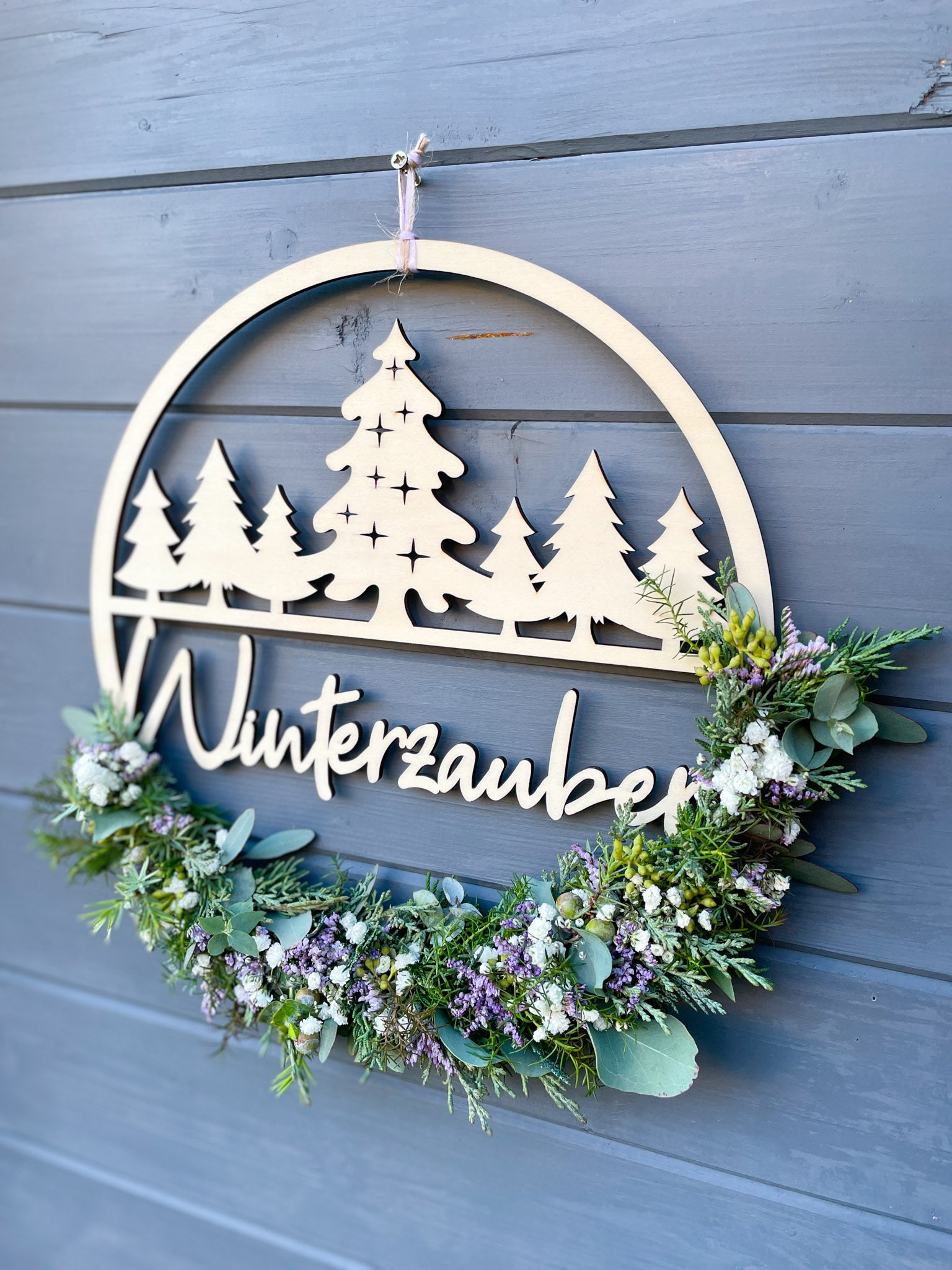 Holzloop Winterzauber mit Schriftzug, bestückt mit Koniferengrün, Eukalyptus und Strandflieder Limonium in Lila, winterliche Dekoration, Ø 25–30 cm, vor grauer Wand