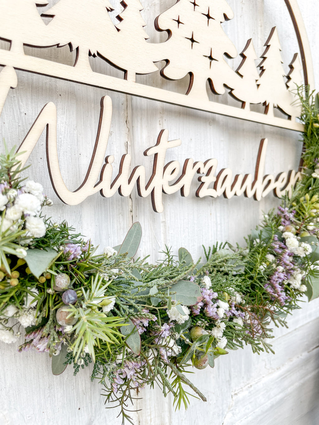 Detail Holzloop Winterzauber, Schriftzug, winterliche Materialien aus Eukalyptus, Koniferengrün und Strandflieder, vor weißer Wand