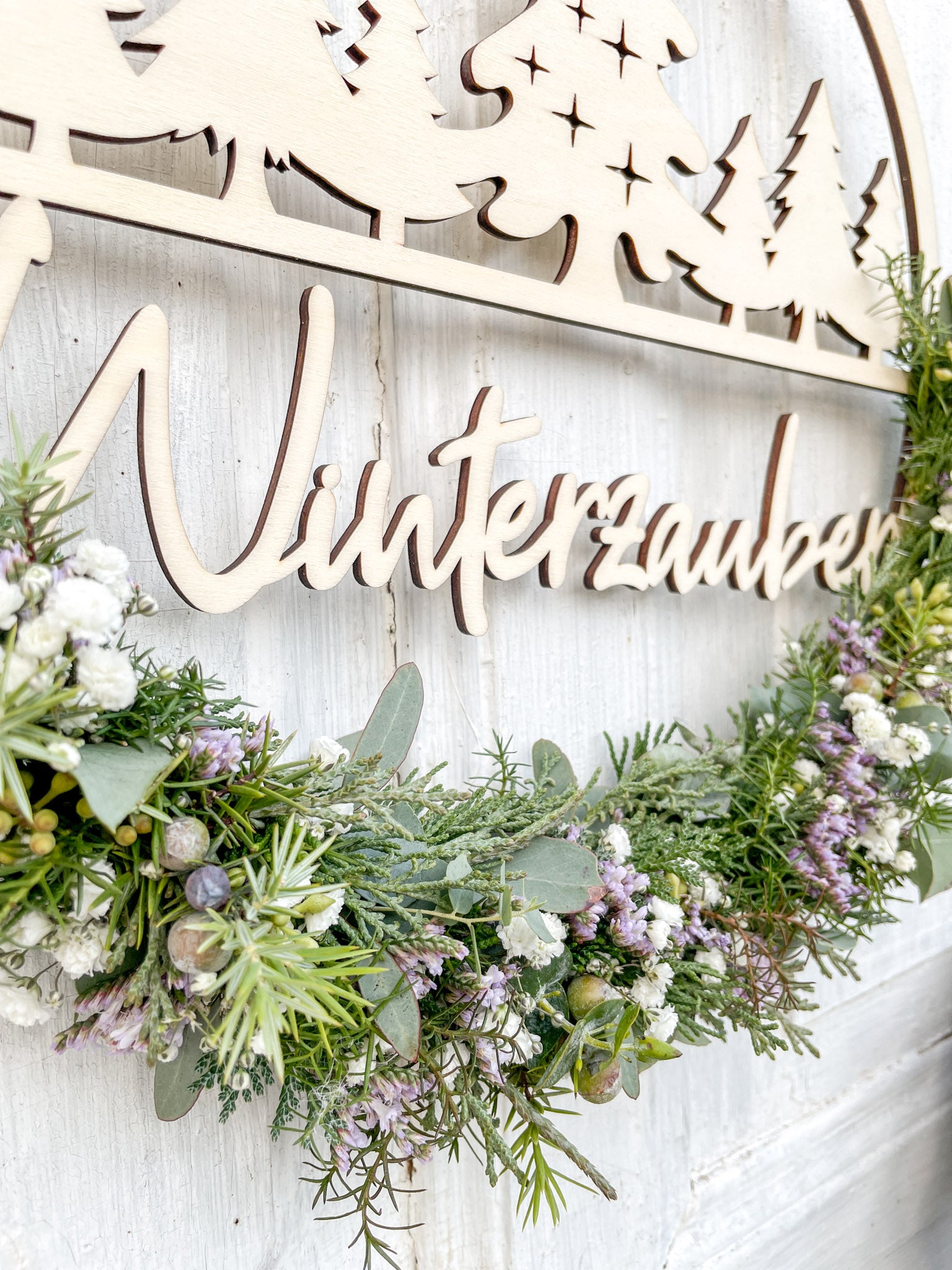 Detail Holzloop Winterzauber, Schriftzug, winterliche Materialien aus Eukalyptus, Koniferengrün und Strandflieder, vor weißer Wand