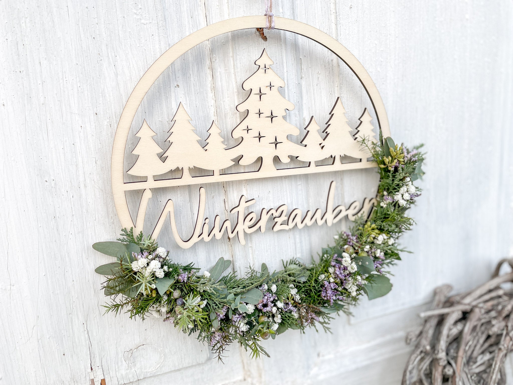 Winterlicher Holzloop Winterzauber, Schriftzug aus Holz, bestückt mit Eukalyptus, Koniferengrün und Lila Strandflieder, Ø 25–30 cm, vor weißer Wand