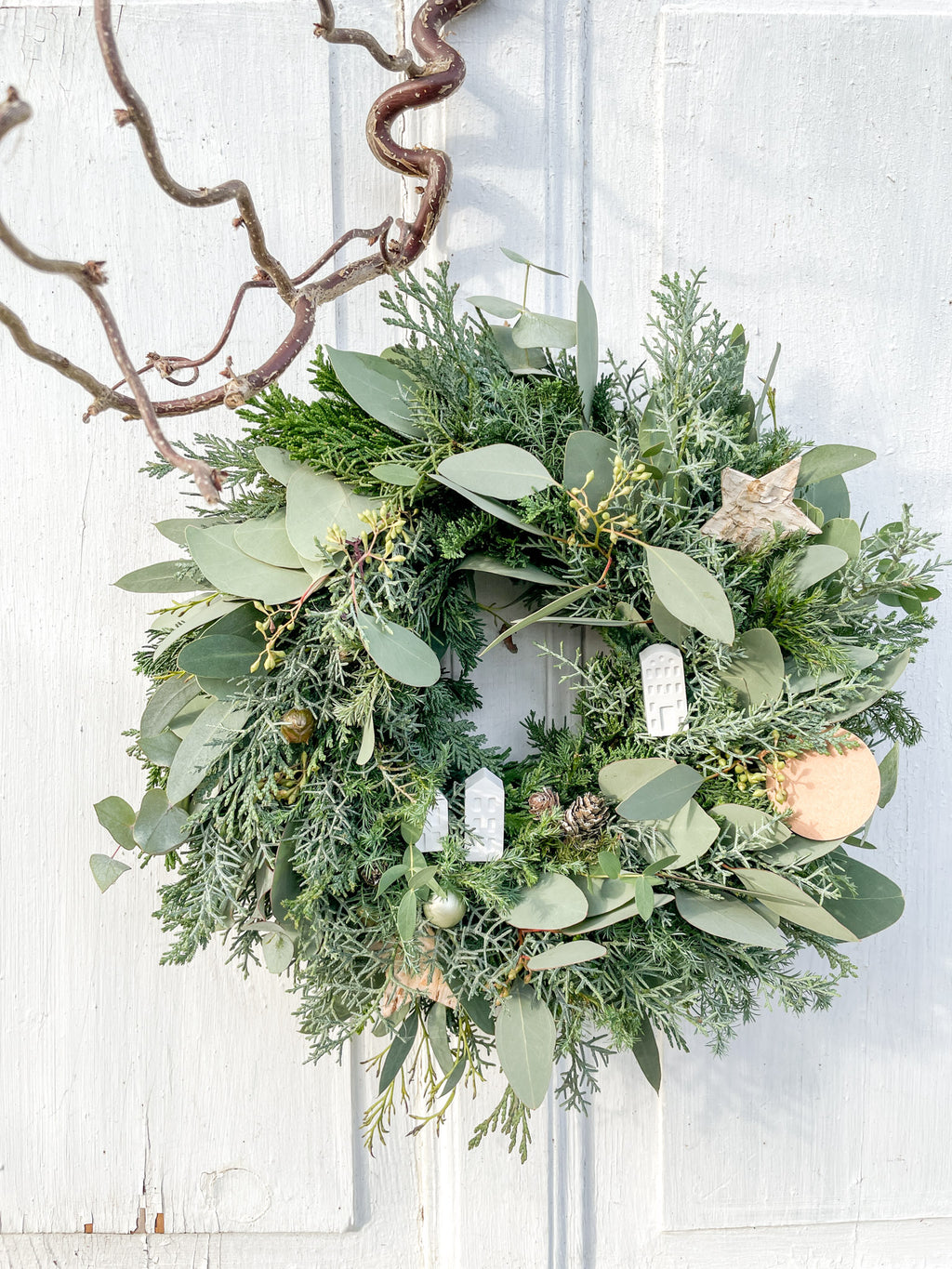 Winterkranz ‚Frosty Village‘ in Hochformatansicht, Tannen- und Koniferengrün mit Eukalyptus und kleinen weißen Häusern, dekorativ arrangiert als Tür- oder Tischkranz