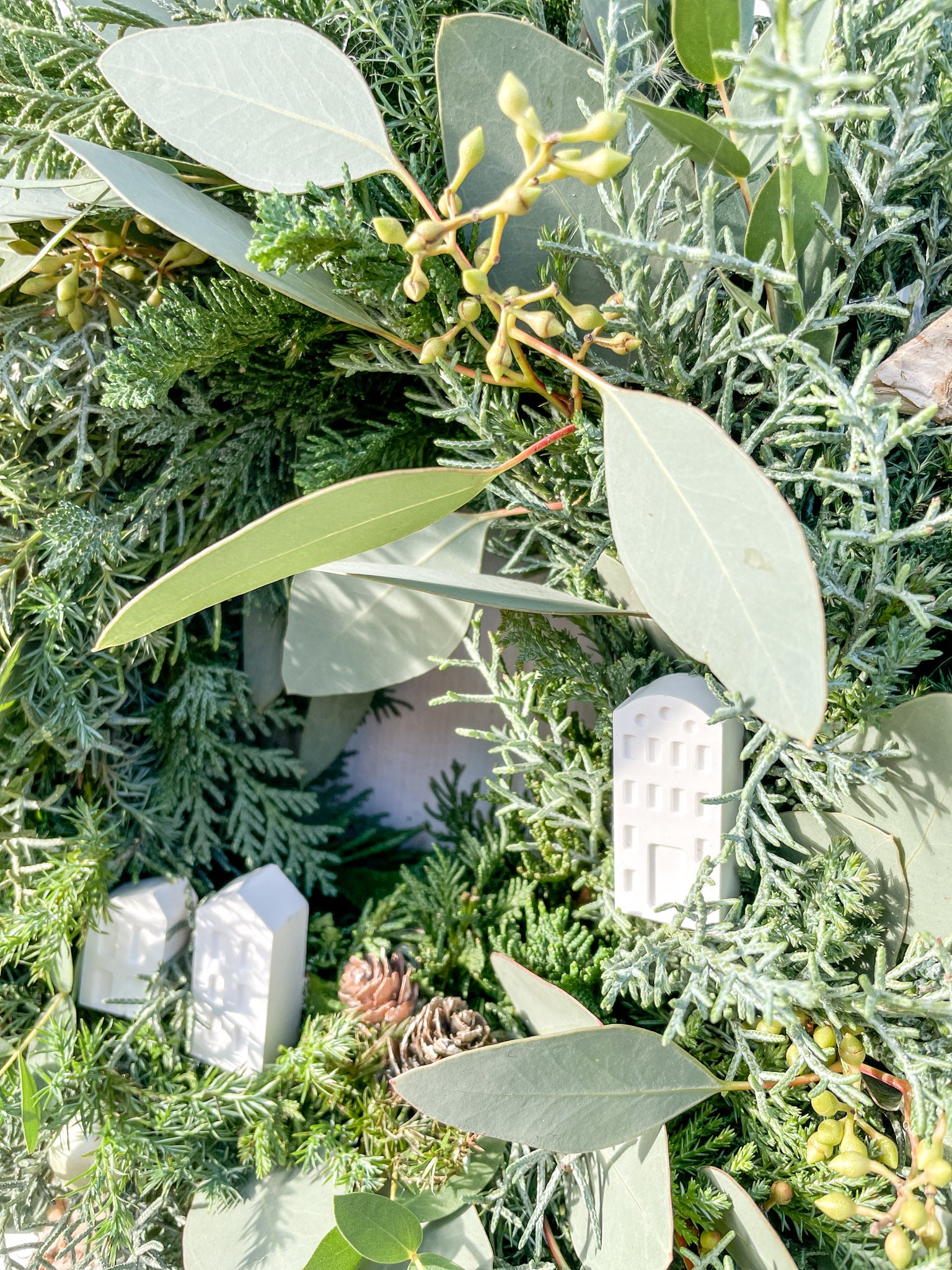 Nahaufnahme der weißen Resin-Häuser auf Winterkranz ‚Frosty Village‘, filigrane Dorfoptik auf Tannen- und Koniferengrün, winterliche Dekoration im minimalistischen Stil